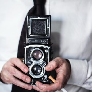 stampa uomo con Rolleiflex muru muru fiorsa primo piano di una macchina fotografica Rolleiflex, tenuta in mano da un uomo che tiene una sigaretta tra le mani, vestito con una camicia bianca e cravatta nera