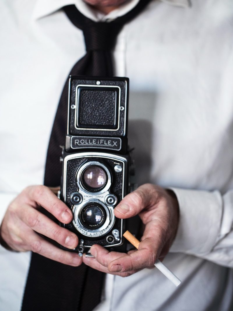 stampa uomo con Rolleiflex muru muru fiorsa primo piano di una macchina fotografica Rolleiflex, tenuta in mano da un uomo che tiene una sigaretta tra le mani, vestito con una camicia bianca e cravatta nera