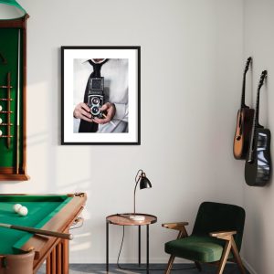 stampa uomo con Rolleiflex muru muru fiorsa ambientata una grande foto con un uomo che tiene in mano una vecchia macchina fotografica Rolleiflex, appesa in una sala giochi, vicino a delle chitarre e a un biliardo