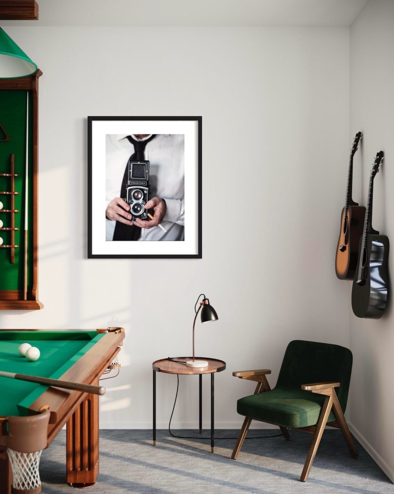 stampa uomo con Rolleiflex muru muru fiorsa ambientata una grande foto con un uomo che tiene in mano una vecchia macchina fotografica Rolleiflex, appesa in una sala giochi, vicino a delle chitarre e a un biliardo