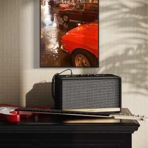 stampa incorniciata di un raduno di auto d'epoca in una città con strade bagnate dalla pioggia, appesa sopra un tavolo su cui è poggiata una chitarra